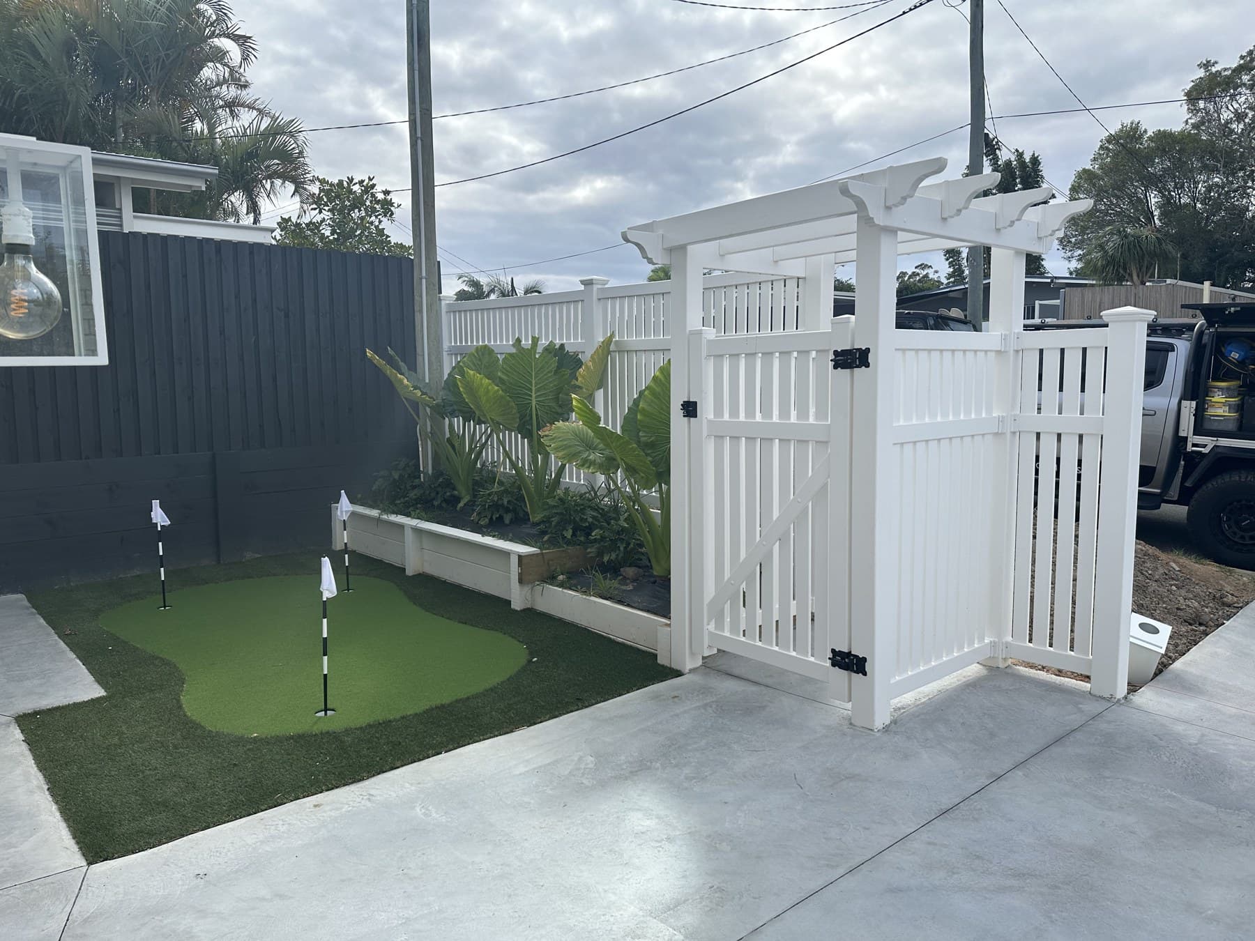 White PVC pedestrian gate with an arbour top, set into a matching PVC fence beside a backyard putting green