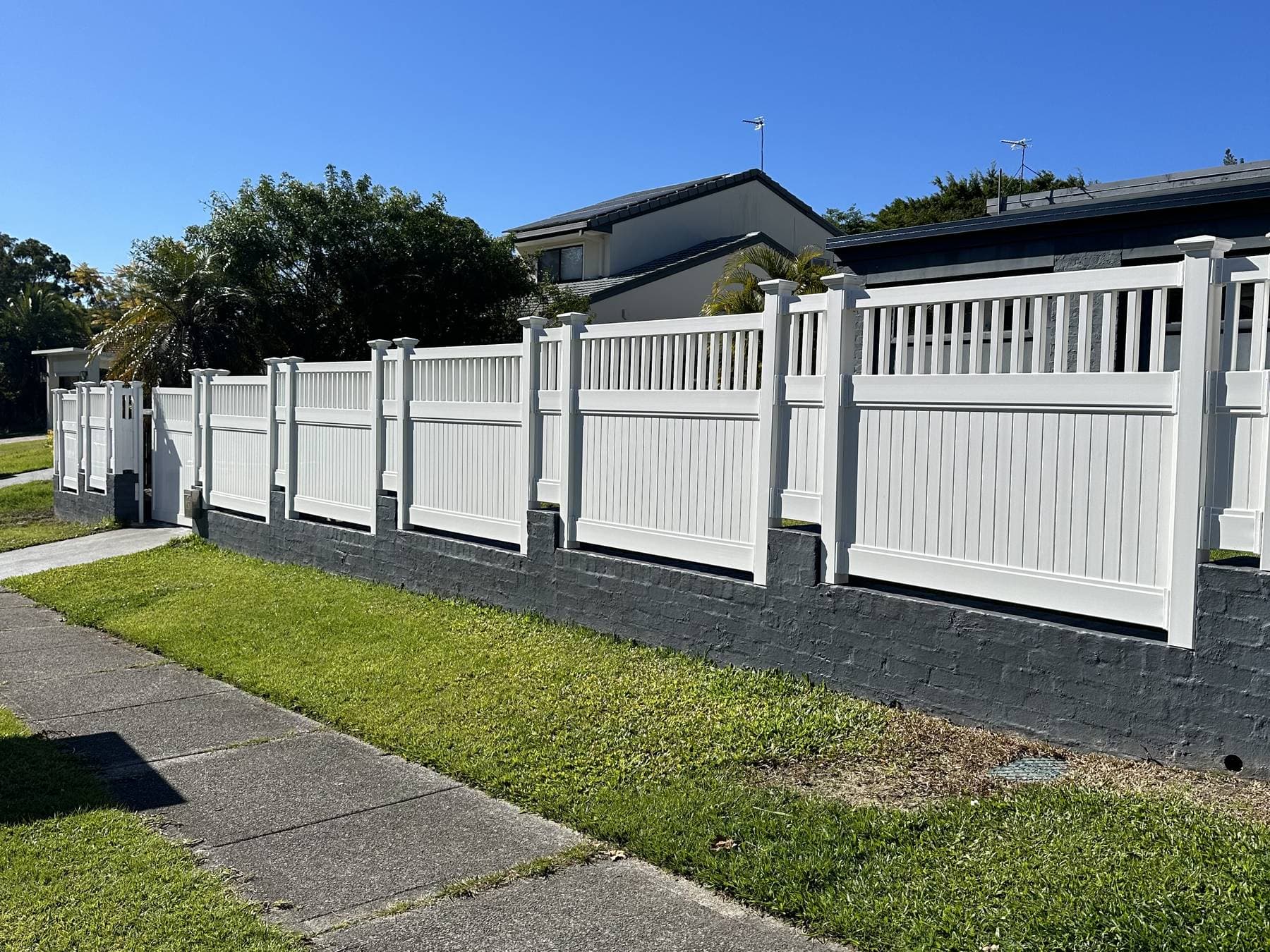 White PVC fence with a slatted top section on a charcoal block retaining wall, running along a suburban footpath