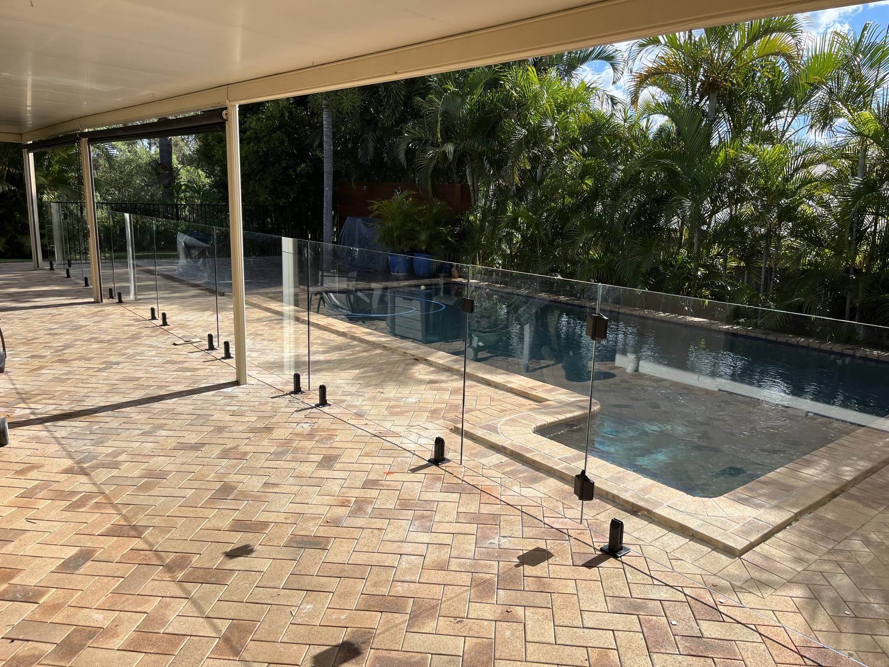 Frameless glass pool fencing on stainless spigots around a pool, under a patio roof with palms behind